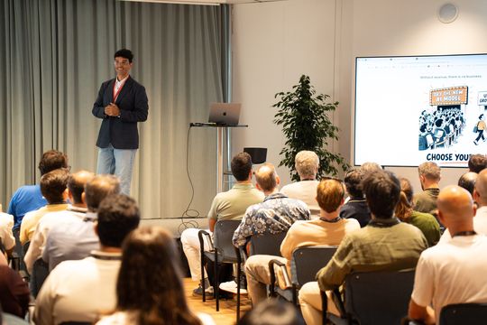 Sami on stage next to the “Choose your queue” slide, room listening