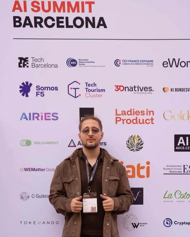 Radhi Badache with his attendee badge, in front of the AI Summit Barcelona community partners wall
